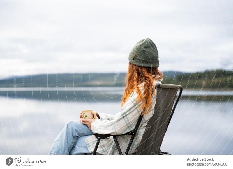 Reflective woman savoring peace by a serene lake calm nature outdoors camping red hair beanie mug chair reflection tranquility solitude water relaxation