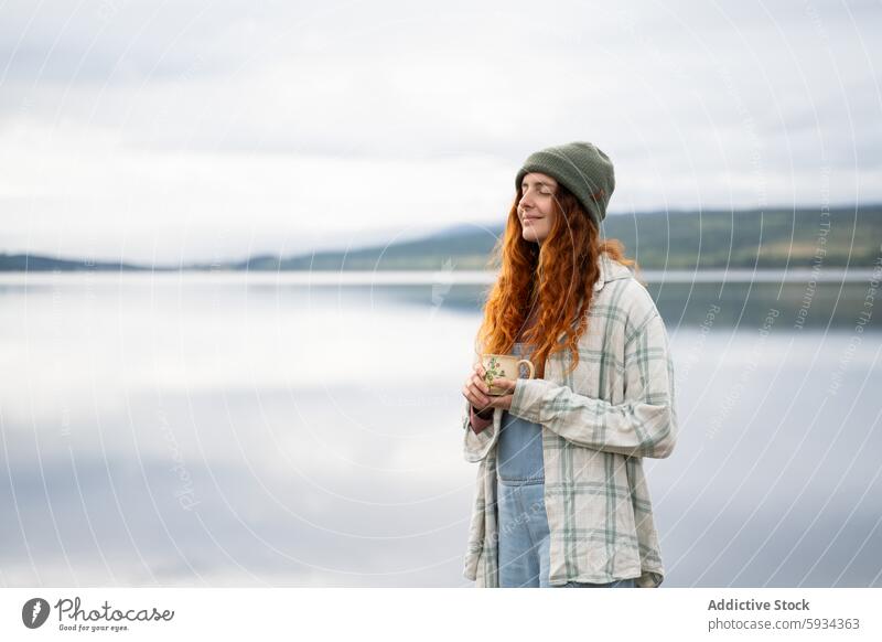 Serene young woman enjoying coffee by the lake morning camping outdoor tranquil serene peaceful redhead beanie plaid shirt nature leisure relaxation lakeside