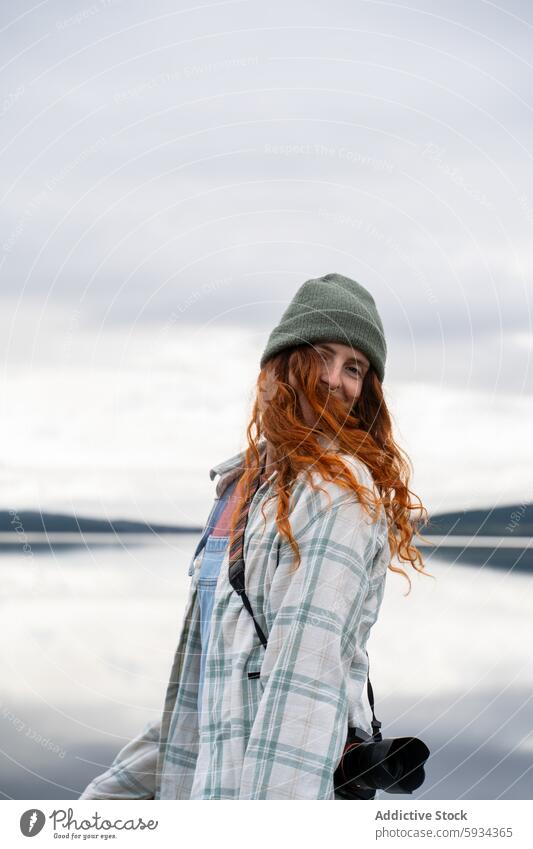 Young woman enjoying a lakeside camping trip outdoors beanie smile nature camera serene cloudy red hair plaid shirt photography leisure activity adventure