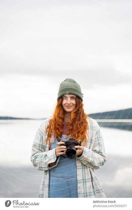 Red-haired woman with camera enjoying lakeside camping red hair outdoor beanie serene nature calm photography leisure activity green cheerful youth vibrant