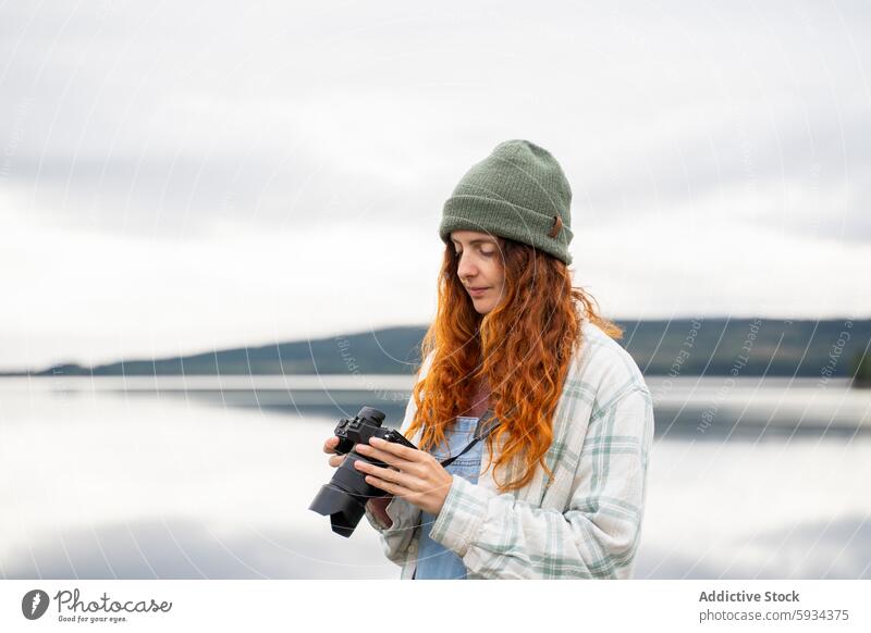 Young woman using camera by a serene lake nature outdoor redhead beanie exploring tranquility landscape scenic view water reflection sky cloud serenity peaceful