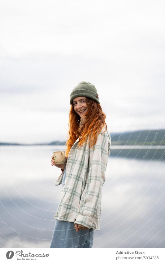 Young woman enjoying a serene lakeside camping trip mug smiling nature outdoor casual tranquility red hair hat beanie shirt plaid style leisure relaxation water