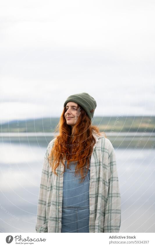 Young woman enjoying serene nature by the lakeside camping tranquility red hair beanie plaid shirt denim dress outdoors leisure landscape water reflection calm