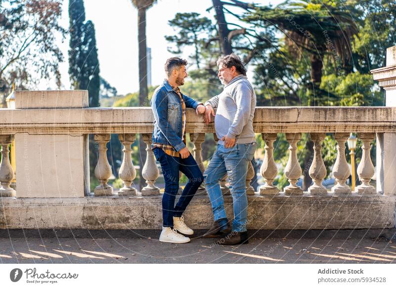 Gay couple enjoying a sunny day at a historic balustrade gay men love park trees architecture companionship leaning happy denim jacket sweater jeans sneaker