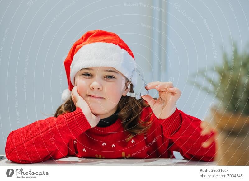 Little girl in festive outfit for Christmas, holding a Christmas cookie cutter christmas baking holiday santa hat sweater kitchen cooking happy joyous seasonal