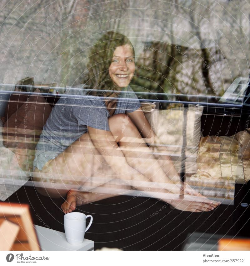 young woman behind a window sits leggy and barefoot on the sofa Young woman Youth (Young adults) Body Legs 18 - 30 years Adults T-shirt Barefoot Brunette