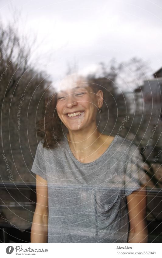 young laughing woman behind a window pane Living room Young woman Youth (Young adults) Face pit 18 - 30 years Adults T-shirt Brunette Long-haired Curl Movement